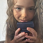 Nevaeh joined the competition — help win amazing prizes! child, curly_hair, face, hands, blue_object, playing, close_up, portrait, indoor, curious, young, person, long_hair, smiling, casual, expression, holding, blurry_background, light, cute