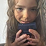 child, curly_hair, face, hands, blue_object, playing, close_up, portrait, indoor, curious, young, person, long_hair, smiling, casual, expression, holding, blurry_background, light, cute