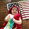 baby, infant, child, smiling, happy, portrait, red_towel, wrapped, sitting, lying_down, package, hands, face, eyes, indoor, wooden_floor, american_flag, rug, home, photography