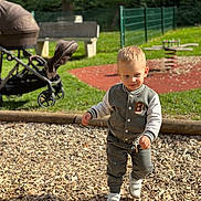 Aaron a rejoint le concours — aidez-le/la à gagner de superbes lots ! toddler, child, playground, wood_chips, stroller, bench, grass, fence, spring, sunlight, jacket, pants, sneakers, outdoor, walking, park, daytime, person, cute, young