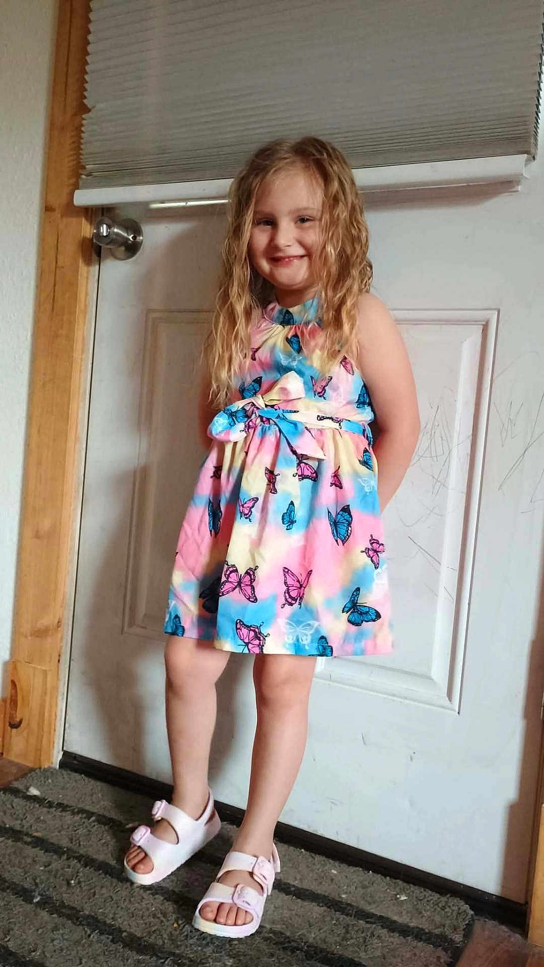 Davina joined the competition — help win amazing prizes! child, girl, smiling, dress, butterfly_pattern, sandals, curly_hair, indoor, door, carpet, wood_frame, happy, standing, portrait, footwear, person, young, casual, colorful, home