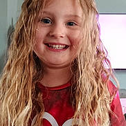 Davina is registered to the contest to win money with this photo: child, smiling, blonde_hair, curly_hair, red_shirt, heart_design, indoor, television, happy, portrait, person, casual_clothing, young, face, eyes, long_hair, light_skin, home, expression, closeup