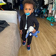 Jahylan a rejoint le concours — aidez-le/la à gagner de superbes lots ! child, toddler, pacifier, stuffed_toy, indoor, flooring, clothing, shoes, toy, living_room, furniture, couch, person, hair, face, expression, standing, wood_floor, black_outfit, blue