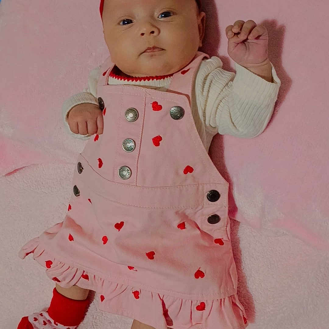 Dynasty is registered to the contest to win money with this photo: baby, bed, buttons, clothing, cozy, cute, eyes_open, face, girl, hand, headband, heart_pattern, infant, lying_down, newborn, pink_blanket, pink_dress, portrait, socks, soft_texture