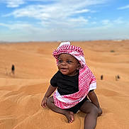 Ysmaël a rejoint le concours — aidez-le/la à gagner de superbes lots ! baby, child, desert, sand, sky, clouds, headwear, keffiyeh, black_clothing, smile, sitting, barefoot, outdoor, sunny, happy, portrait, nature, people, infant, young_child