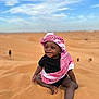 baby, child, desert, sand, sky, clouds, headwear, keffiyeh, black_clothing, smile, sitting, barefoot, outdoor, sunny, happy, portrait, nature, people, infant, young_child