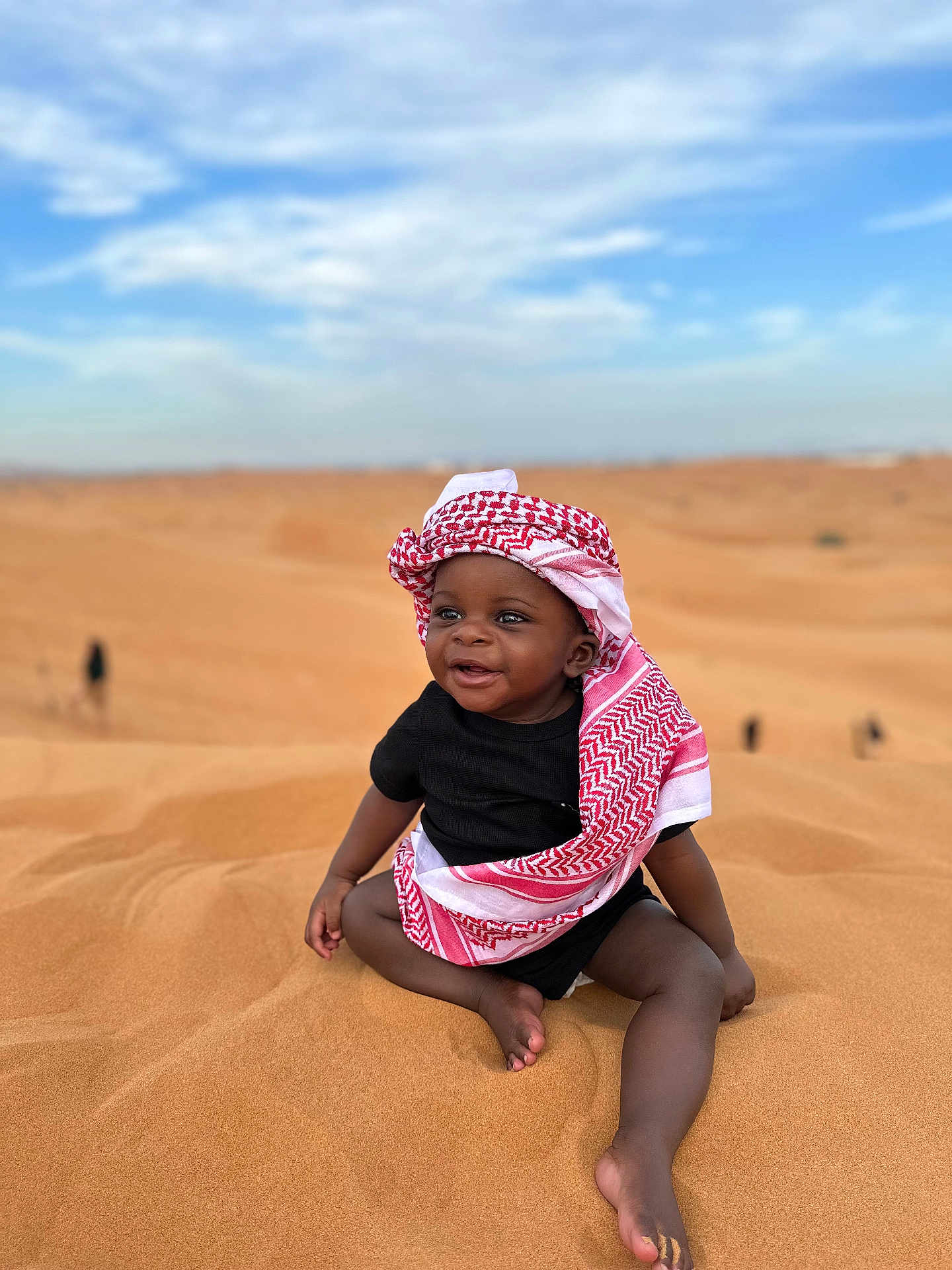 Ysmaël a rejoint le concours — aidez-le/la à gagner de superbes lots ! baby, child, desert, sand, sky, clouds, headwear, keffiyeh, black_clothing, smile, sitting, barefoot, outdoor, sunny, happy, portrait, nature, people, infant, young_child