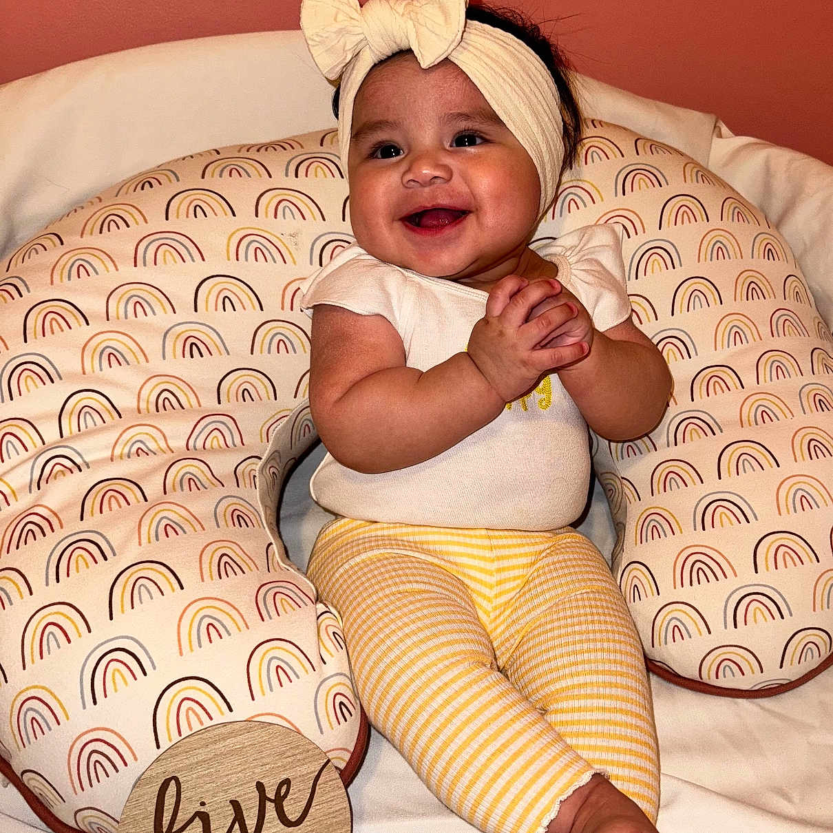 Madelyn joined the competition — help win amazing prizes! baby, child, clothing, cute, feet, five_months, hands, happy, head, headband, indoor, infant, pillow, portrait, rainbow_pattern, sitting, smiling, white_background, wooden_sign, yellow_pants