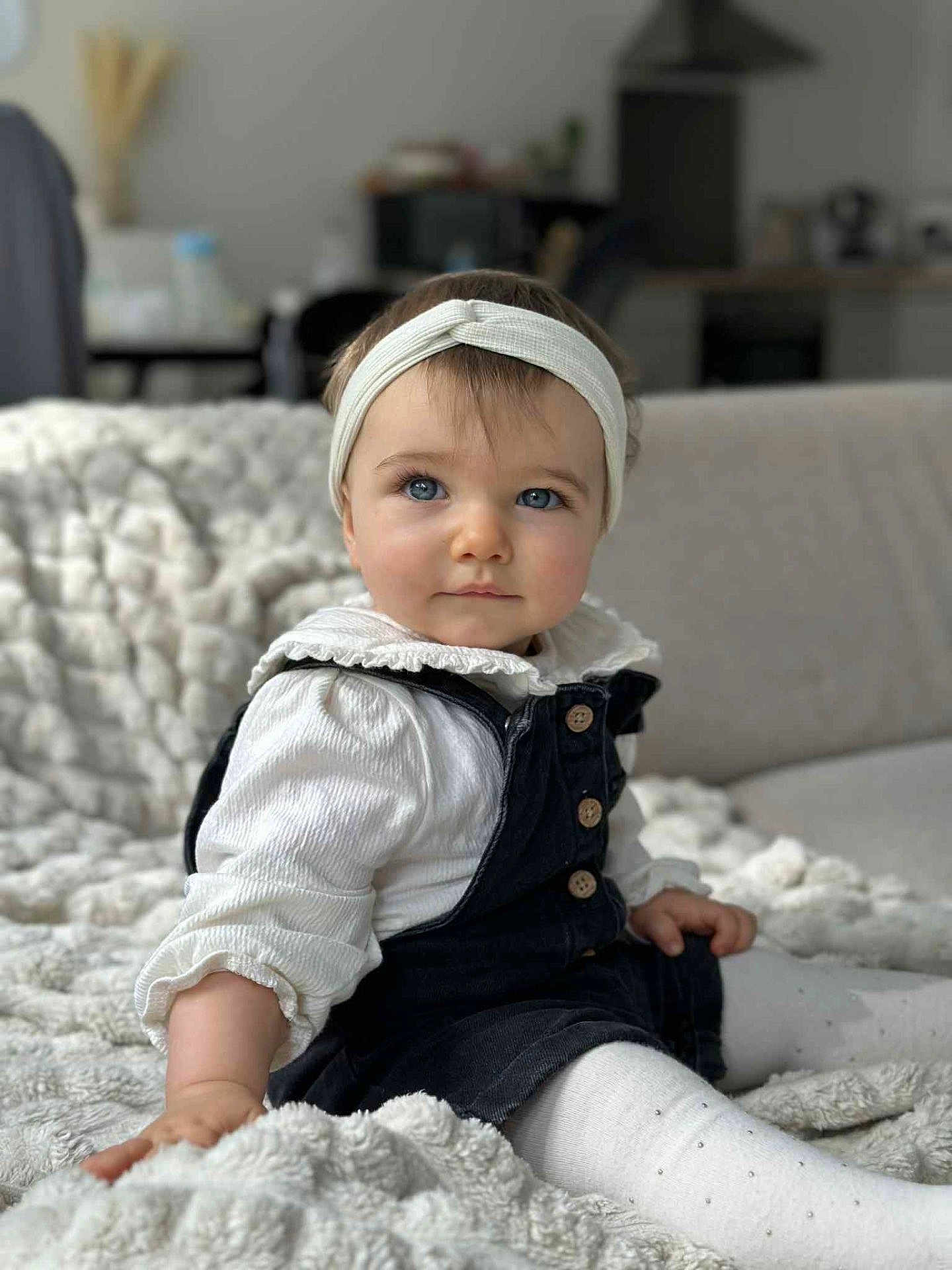Kiara a rejoint le concours — aidez-le/la à gagner de superbes lots ! baby, child, headband, blue_eyes, white_shirt, dark_overalls, blanket, cozy, indoor, sitting, portrait, cute, soft_lighting, infant, young_child, comfortable, curious, homedecor, texture, casual_clothing