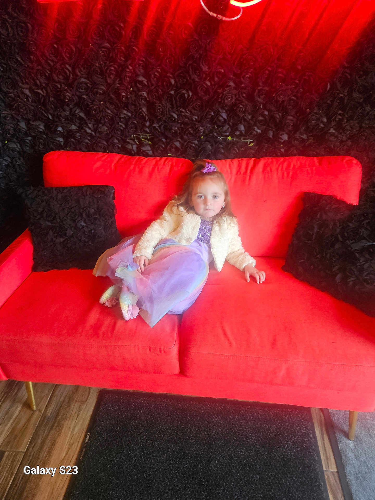 Ariel joined the competition — help win amazing prizes! child, girl, couch, red_couch, purple_dress, white_jacket, black_pillows, floral_wall, indoor, carpet, wood_floor, portrait, seated, looking_at_camera, curly_hair, hair_bow, casual_pose, home_interior, furniture, lighting