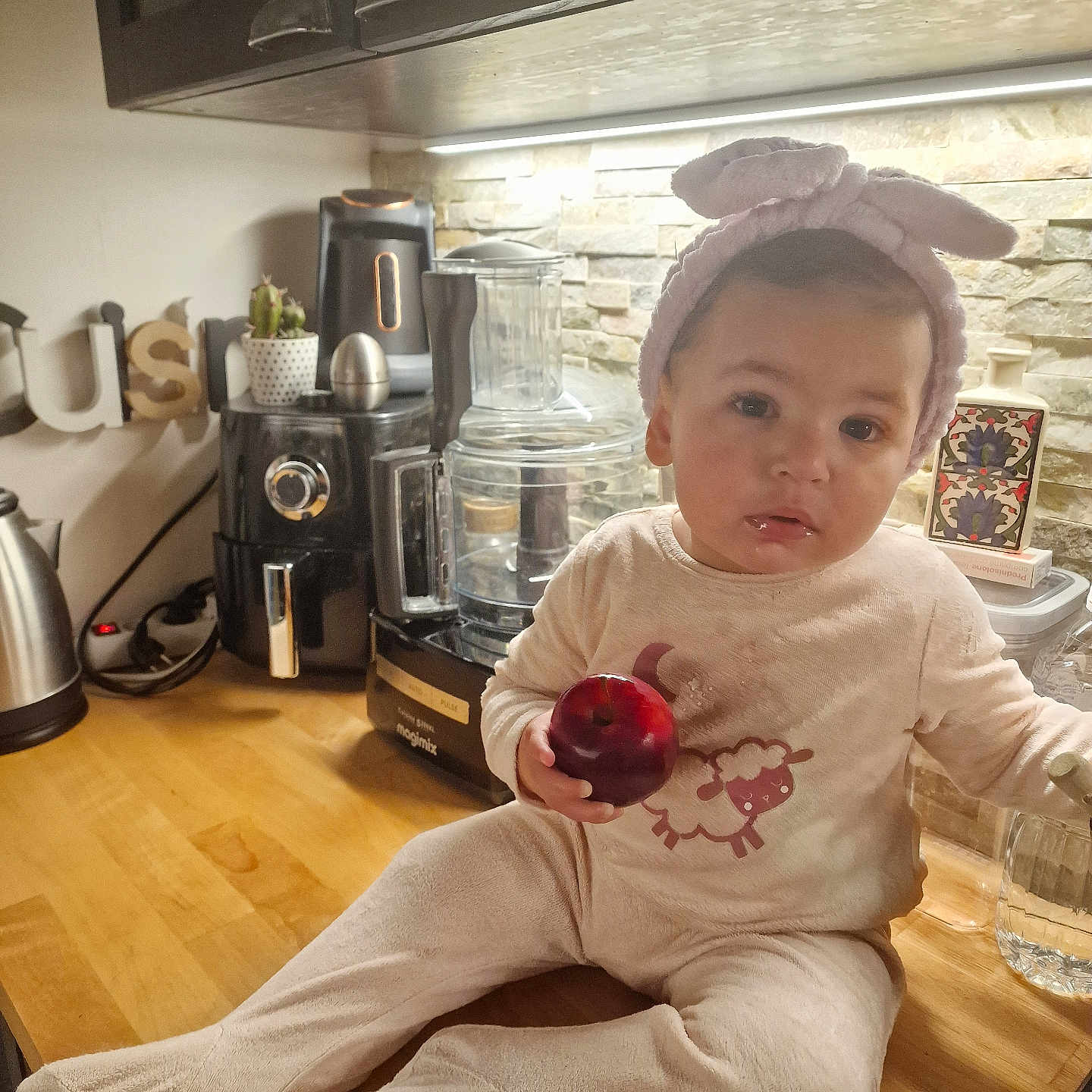 Ilef a rejoint le concours — aidez-le/la à gagner de superbes lots ! apple, appliance, baby, baby_clothing, bunny_ears, child, countertop, cute, food, fruit, headband, home, indoors, kitchen, onesie, person, portrait, sitting, table, warm_lighting