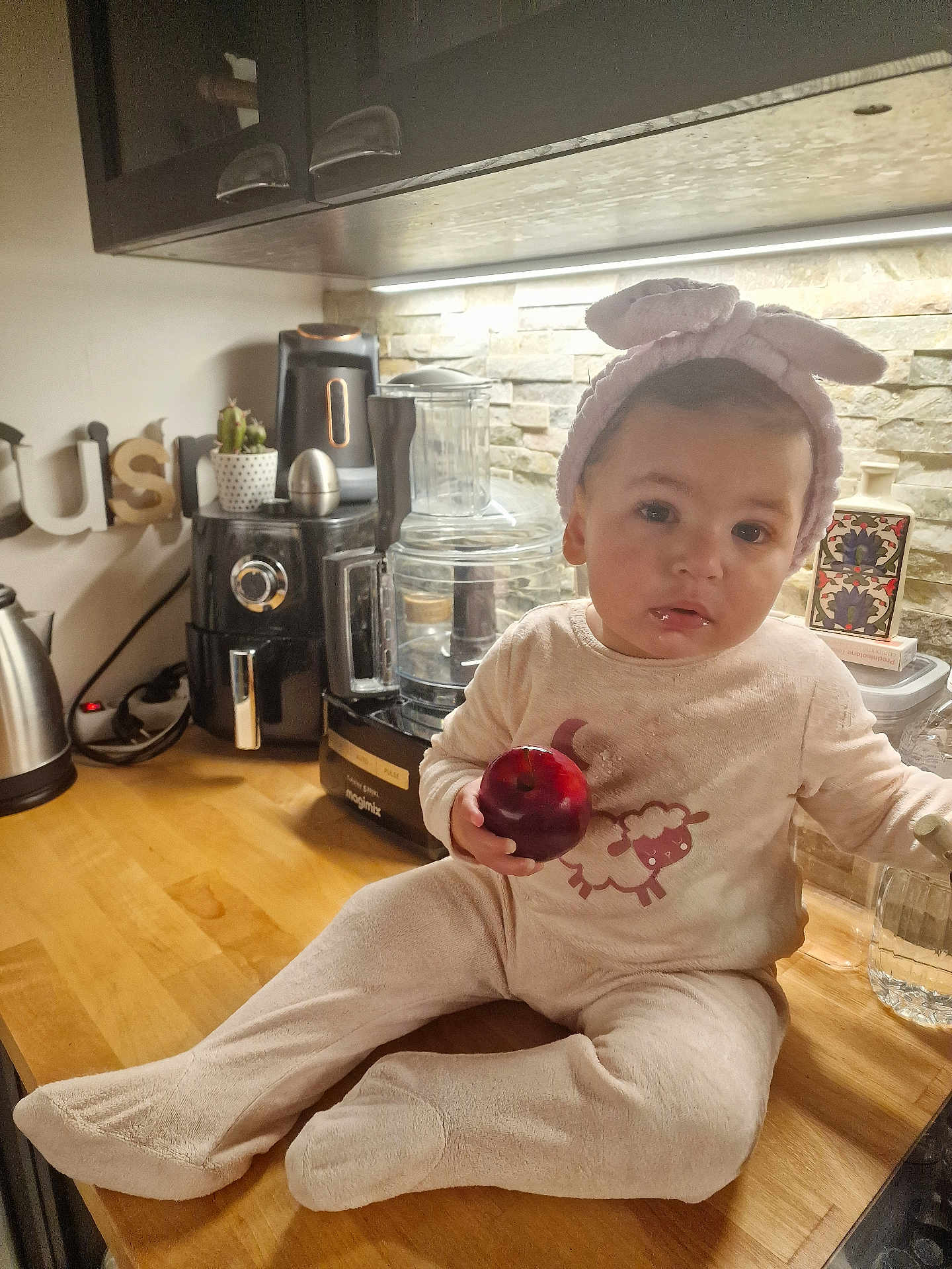 Ilef a rejoint le concours — aidez-le/la à gagner de superbes lots ! baby, child, kitchen, apple, onesie, headband, bunny_ears, countertop, appliance, food, cute, indoors, person, portrait, table, fruit, baby_clothing, sitting, home, warm_lighting