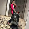 woman, mirror_selfie, dog, stroller, hallway, tiled_floor, pink_shirt, smartphone, long_hair, earbuds, black_pants, sneakers, indoor, pet, pet_stroller, reflection, door, elevator, handbag, casual