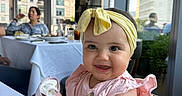 Harlow joined the competition — help win amazing prizes! baby, child, smile, headband, pink_dress, high_chair, pacifier, indoor, restaurant, table, window, cityscape, people, woman, glass, food, furniture, happy, portrait, cute