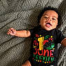 baby, infant, smiling, curly_hair, black_onesie, juneteenth, celebration, text_on_clothing, lying_down, blanket, happy, cute, child, portrait, indoors, skin, hand, face, mouth_open, one_piece_clothing