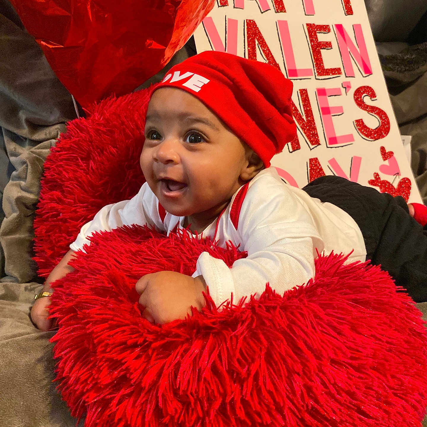 Amir joined the competition — help win amazing prizes! baby, red_hat, red_cushion, valentines_day, balloon, smiling, happy, infant, cute, holiday, celebration, sign, love, indoor, cozy, person, clothing, face, headwear, couch