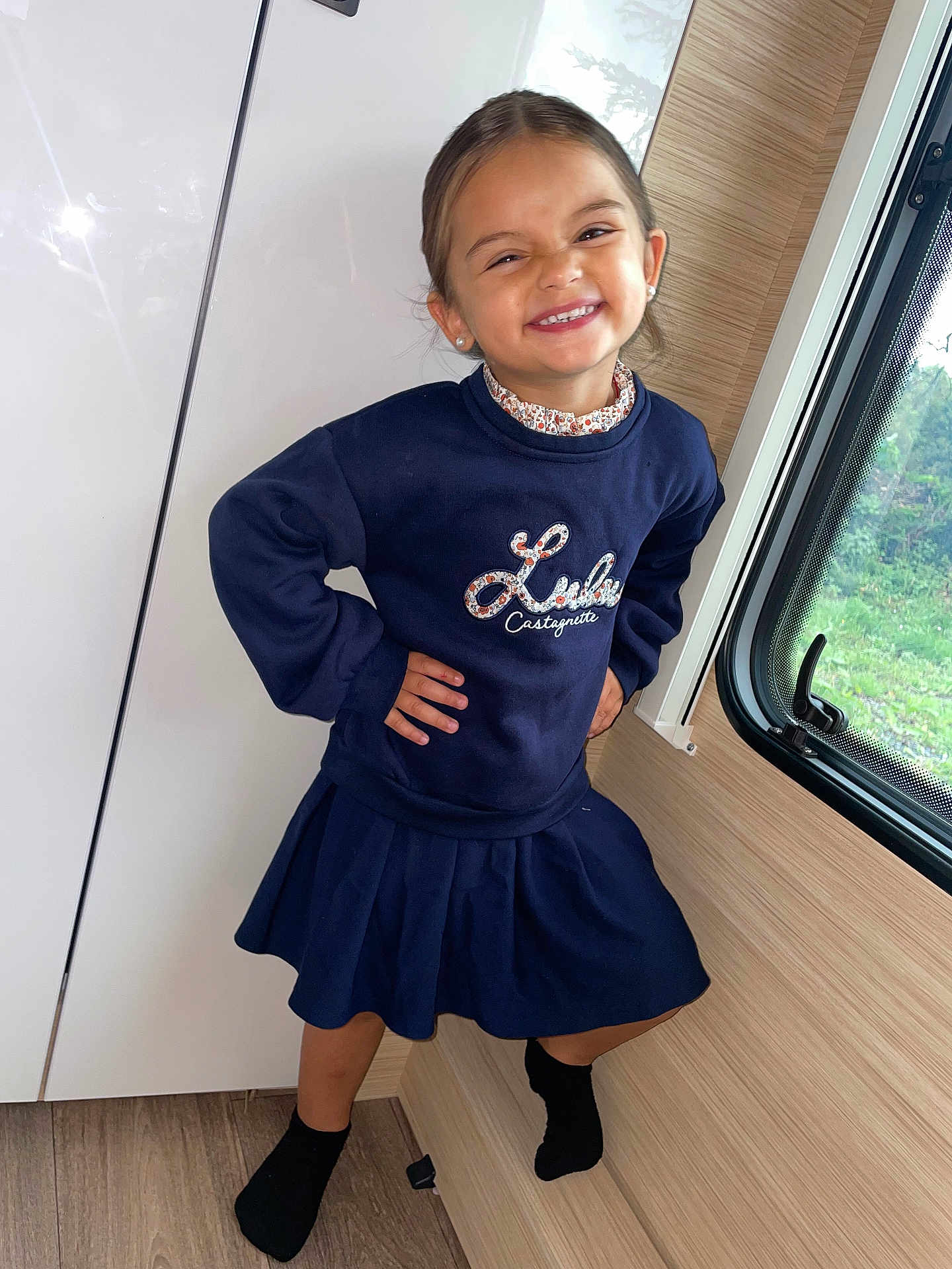 Dailly participe au concours pour gagner de l'argent avec cette photo : child, girl, smile, indoor, window, blue_clothing, skirt, sweatshirt, pose, happy, young, earrings, floor, wooden_floor, cabinet, person, fashion, portrait, feet, sock