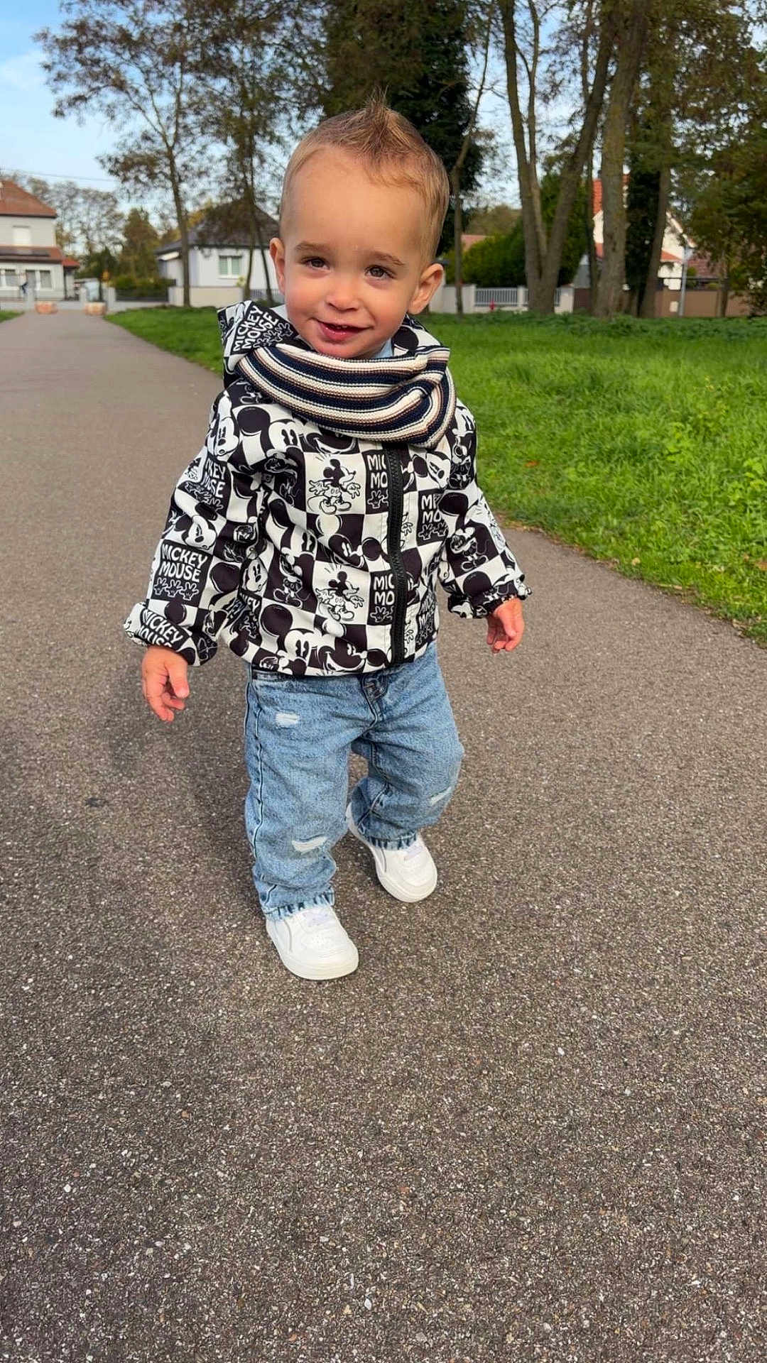 Naël Herrmann a rejoint le concours — aidez-le/la à gagner de superbes lots ! toddler, child, smiling, jacket, mickey_mouse, scarf, jeans, white_shoes, outdoor, sidewalk, trees, grass, houses, autumn, young_child, casual_clothing, portrait, walking, daytime, happy