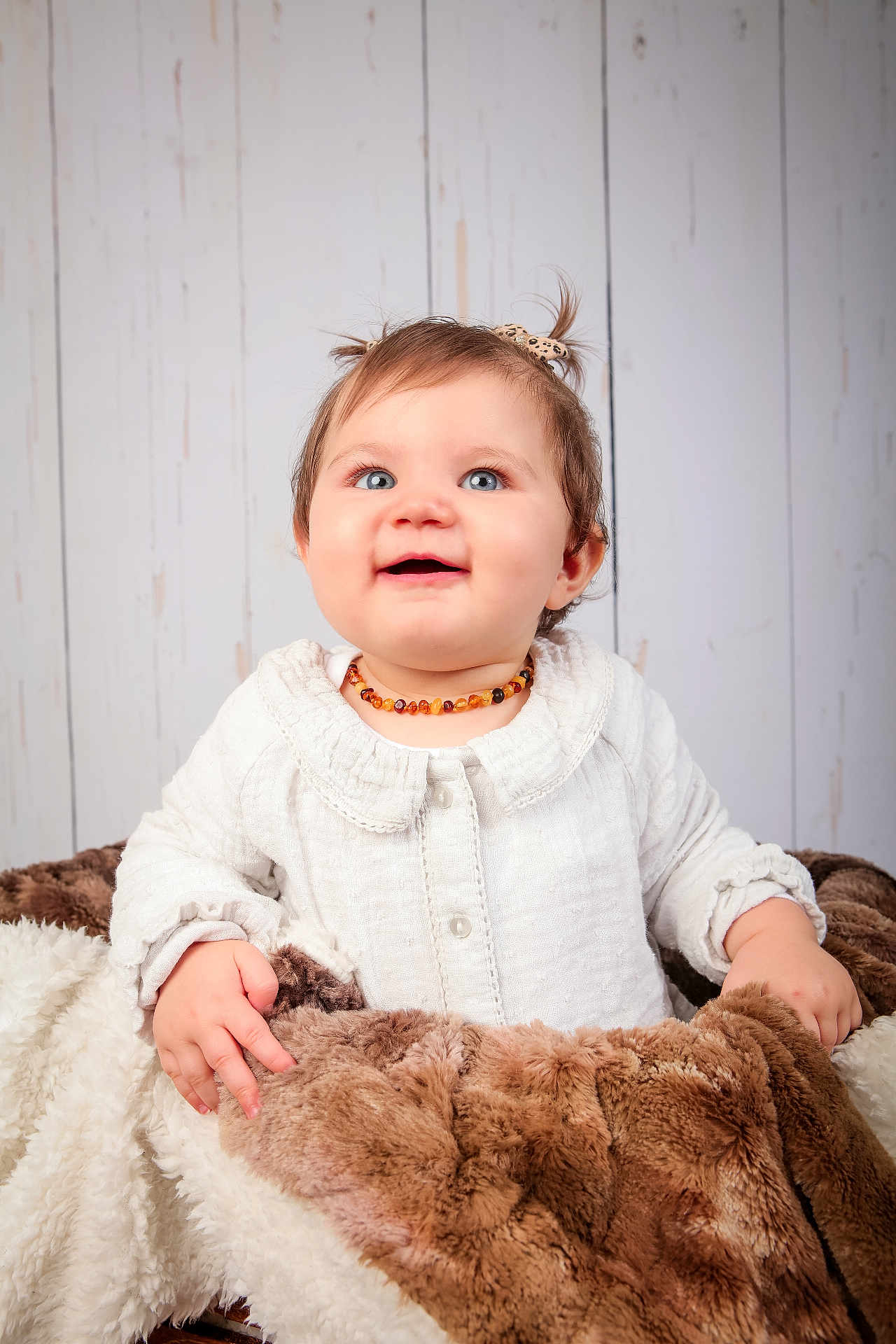 Ambre participe au concours pour gagner de l'argent avec cette photo : baby, child, blue_eyes, pigtails, white_clothing, necklace, blanket, fur_blanket, smiling, happy, portrait, indoor, wooden_background, cute, infant, toddler, hands, face, soft_texture, cozy