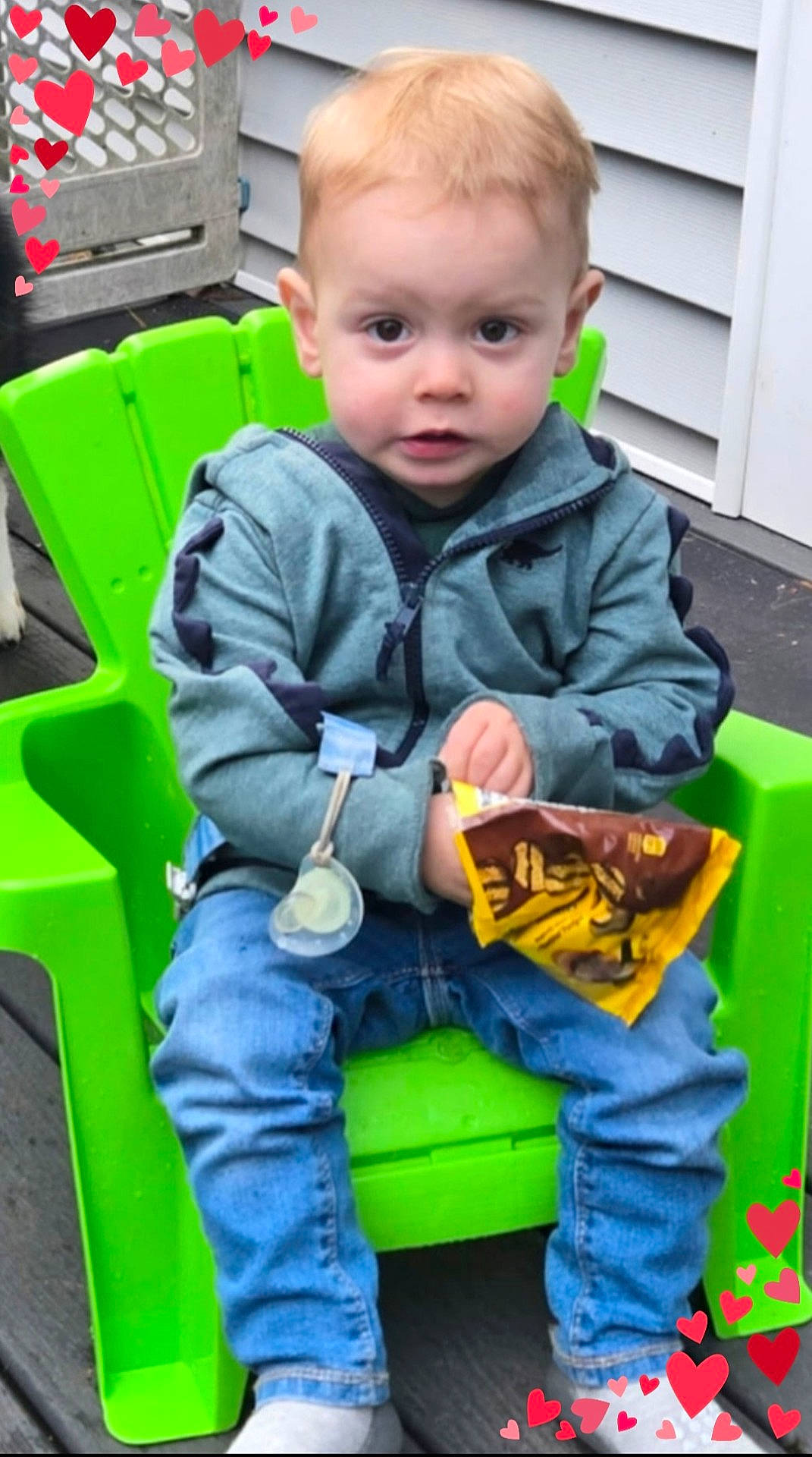 Jaxon is registered to the contest to win money with this photo: casual_clothing, child, cute, daylight, expression, green_chair, heart_graphics, hoodie, jeans, outdoor, pacifier, plastic_chair, porch, red_hearts, sitting, snack, snack_time, toddler, wooden_floor, young_child