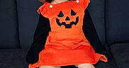Millianna joined the competition — help win amazing prizes! child, toddler, girl, halloween, costume, pumpkin, orange_dress, black_sleeves, sitting, couch, portrait, indoors, brick_wall, brown_hair, curly_hair, barefoot, smiling, face, furniture, holiday