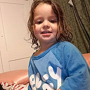 Millianna is registered to the contest to win money with this photo: child, toddler, portrait, smiling, happy, indoor, bedroom, blue_shirt, curly_hair, brown_hair, face, person, sitting, bed, blanket, curtains, door, home, close_up, photography