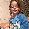 child, toddler, portrait, smiling, happy, indoor, bedroom, blue_shirt, curly_hair, brown_hair, face, person, sitting, bed, blanket, curtains, door, home, close_up, photography