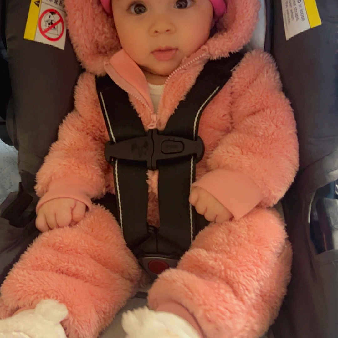 Camila is registered to the contest to win money with this photo: adorable, baby, boots, car_seat, child, cute, expression, fluffy_clothing, hat, indoor, infant, person, pink_onesie, portrait, safety_harness, seated, warm_clothing, wide_eyes, winter_wear, young_child