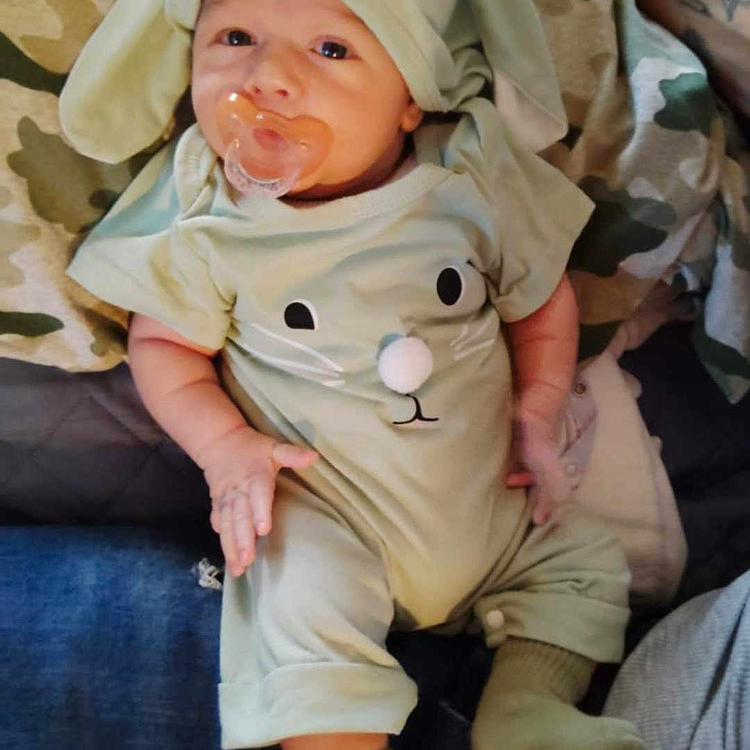Axton is registered to the contest to win money with this photo: baby, bunny_ears, camouflage_blanket, child, costume, couch, cute, fabric, green_clothing, hand, indoors, infant, lap, pacifier, person, portrait, resting, socks, soft_lighting, tattoo