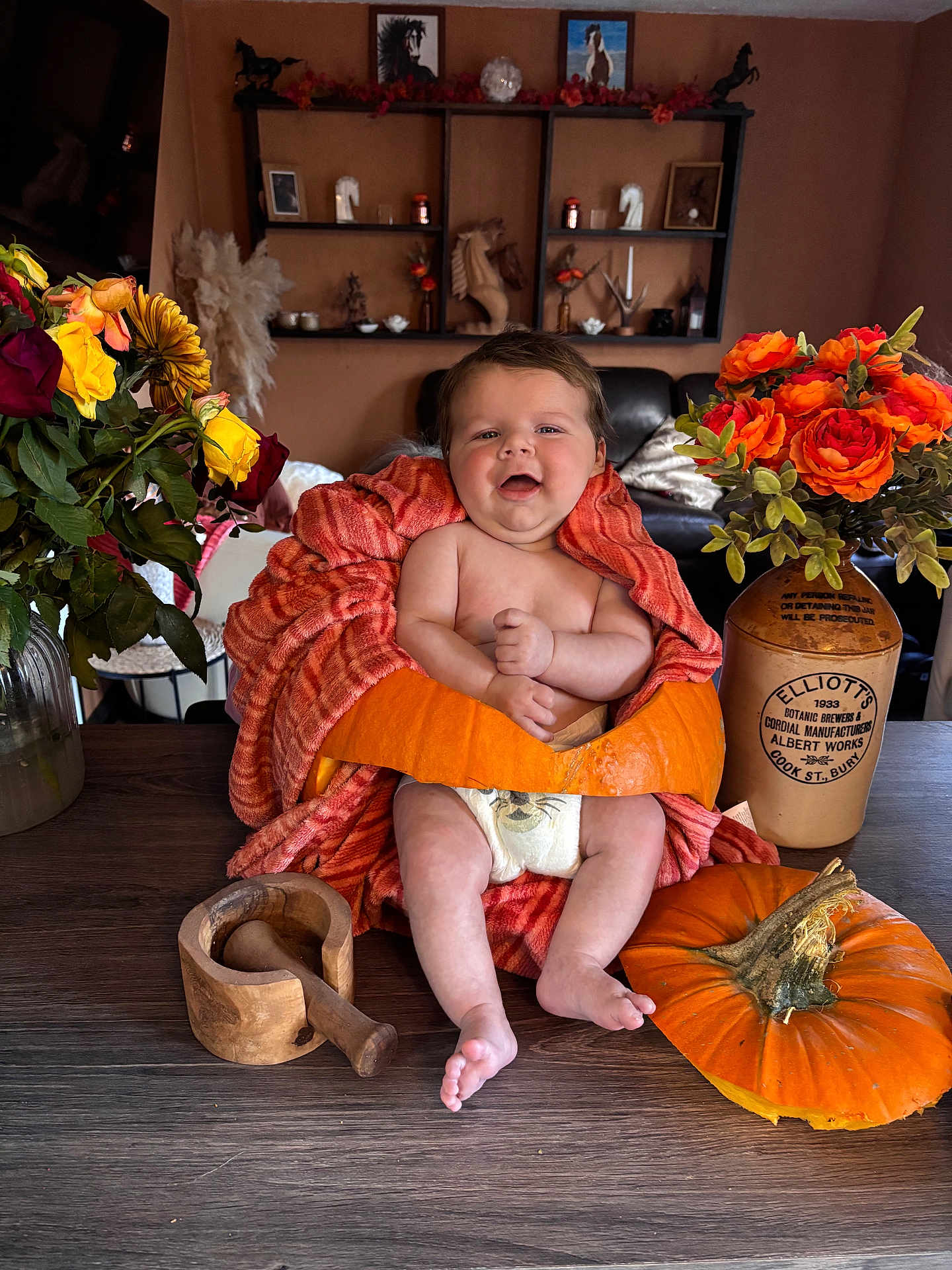 Hudson is registered to the contest to win money with this photo: baby, pumpkin, orange_towel, flowers, vase, wooden_table, mortar_and_pestle, smiling, indoor, decor, diaper, brown_wall, shelf, horse_figurine, plant, pottery, child, happy, portrait, cozy