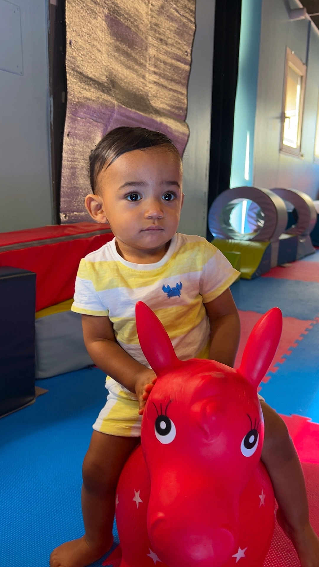 Aïjaz participe au concours pour gagner de l'argent avec cette photo : toddler, child, indoor, play_area, toy, inflatable, red, horse, colorful, mats, padded, shorts, tshirt, young_child, person, play, fun, expression, cute, floor