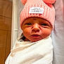 baby, newborn, infant, swaddle, hat, pink_hat, pom_poms, face, eyes, nose, mouth, expression, wrinkled_skin, blanket, bassinet, clothing, portrait, close_up, hospital, adorable