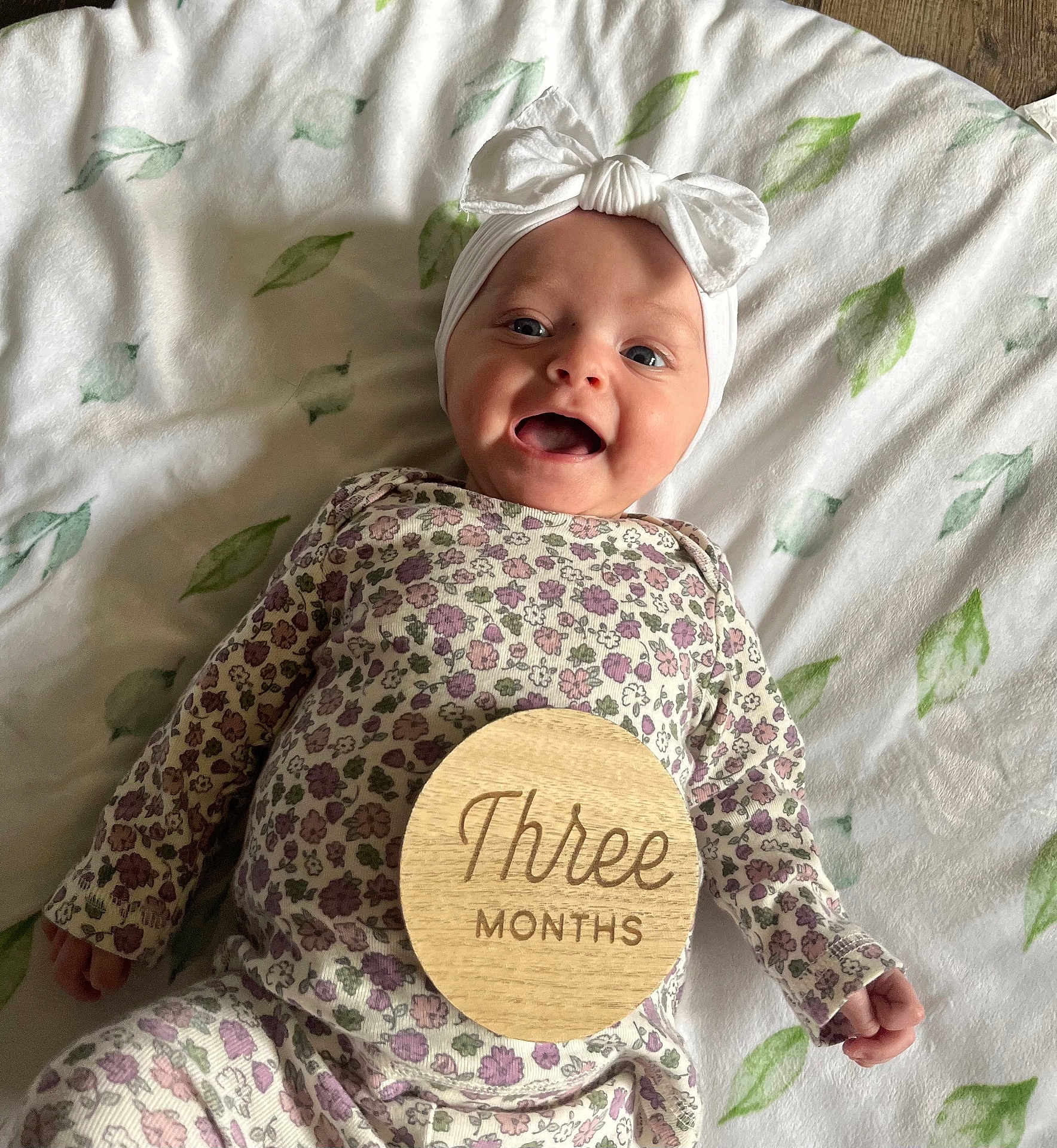 Zarriah is registered to the contest to win money with this photo: baby, infant, smile, open_mouth, headband, bow, wooden_disc, three_months, milestone, floral_outfit, onesie, blanket, leaf_pattern, lying_down, portrait, close_up, happy, soft_lighting, nursery, hand