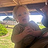 child, toddler, baby, portrait, face, eyes, blond_hair, curious, sitting, being_held, adult, family, outdoor, porch, wooden_ceiling, backyard, house, grass, sunlight, photograph