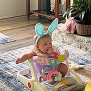 Blakeleigh is registered to the contest to win money with this photo: baby, child, infant, bunny_ears, headband, walker, baby_toy, rainbow_toy, plush_toy, smiling, indoors, living_room, carpet, plant, furniture, sunlight, wood_floor, pink_clothing, sitting, portrait