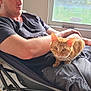 Garfield is registered to the contest to win money with this photo: man, cat, orange_tabby, chair, indoor, window, casual_clothing, relaxed, pet, animal, human, feline, seated, natural_light, home, floor_tiles, short_hair, looking_down, curious, resting