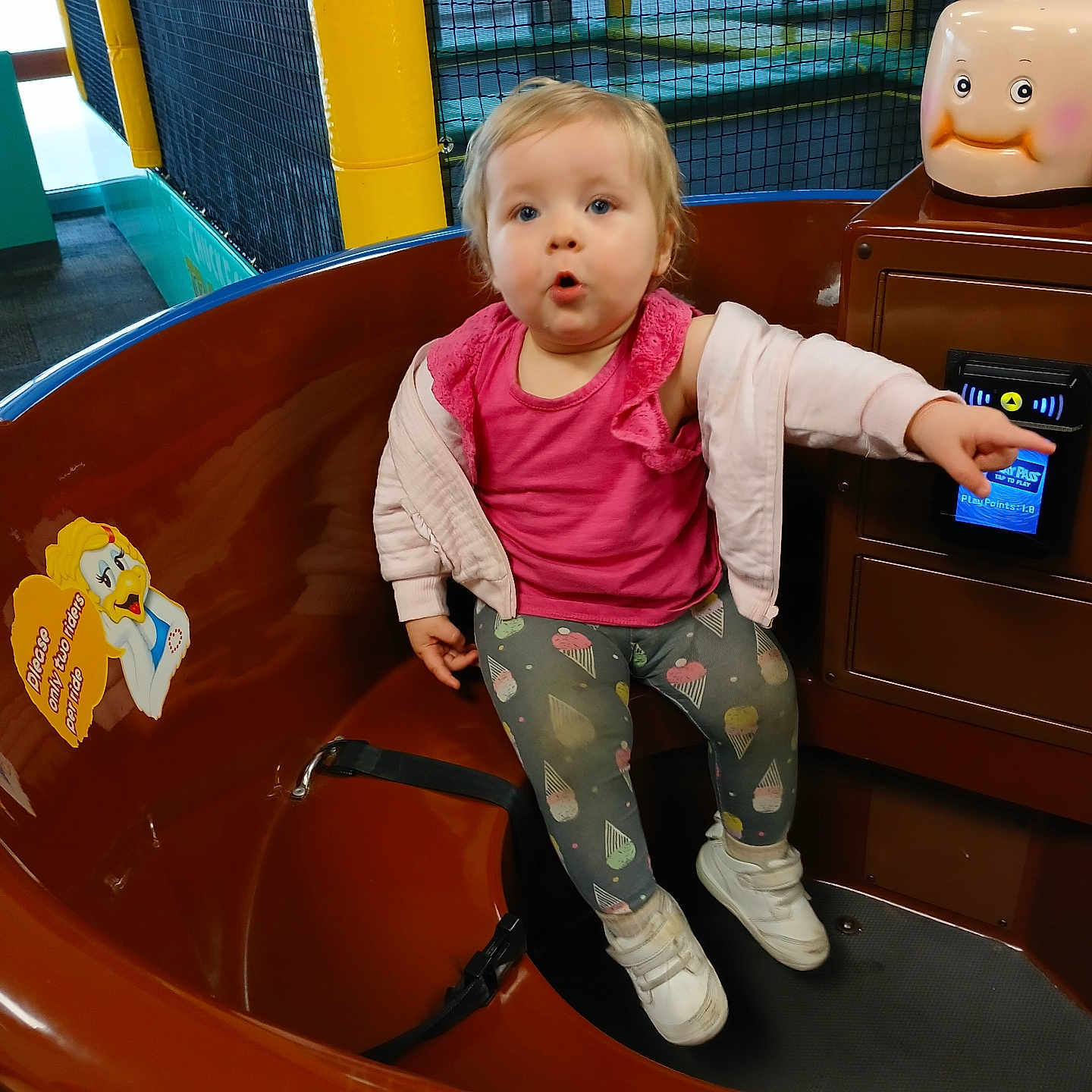 Louann joined the competition — help win amazing prizes! amusement_ride, brown_ride, cartoon_sticker, casual_clothing, child, control_panel, curious_expression, fun, indoor, leggings, netting, pink_top, play_area, playground, pointing, seated, short_hair, toddler, white_shoes, yellow_poles
