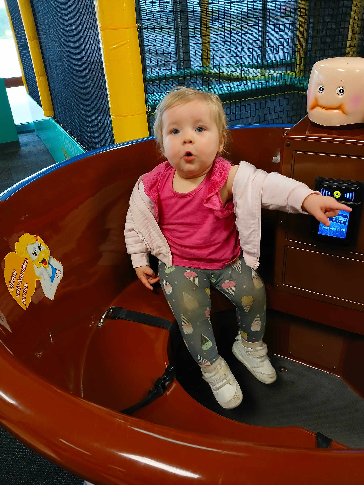 Louann joined the competition — help win amazing prizes! toddler, child, amusement_ride, pink_top, leggings, white_shoes, indoor, play_area, pointing, brown_ride, cartoon_sticker, curious_expression, control_panel, yellow_poles, netting, casual_clothing, short_hair, fun, seated, playground