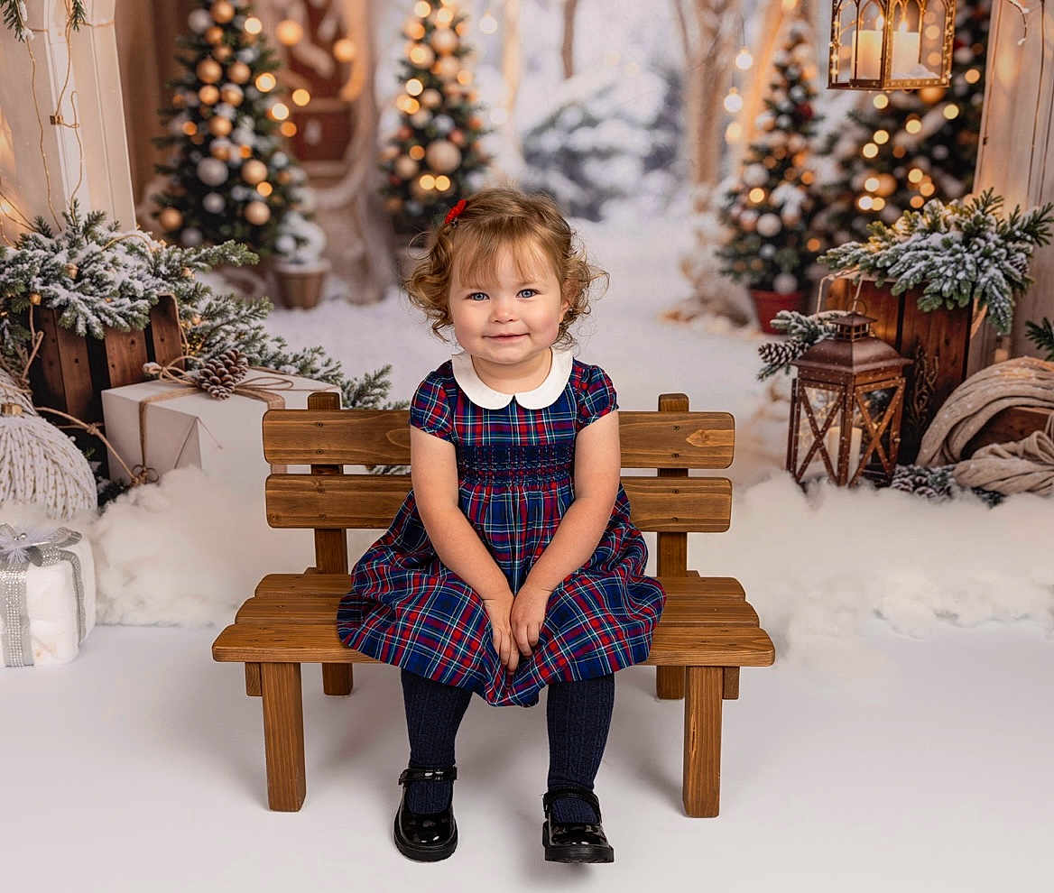 Mimi joined the competition — help win amazing prizes! toddler, child, girl, plaid_dress, wooden_bench, curly_hair, red_bow, black_shoes, christmas_tree, holiday_decor, wrapped_presents, lantern, winter_theme, snow_effect, festive, smiling, indoors, portrait, studio_photo, cozy
