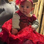 Lilith is registered to the contest to win money with this photo: baby, child, red_dress, headband, adult, indoor, person, smile, curious, holding, wooden_frame, unfinished_wall, long_sleeve, face, closeup, portrait, cute, fashion, female, celebration