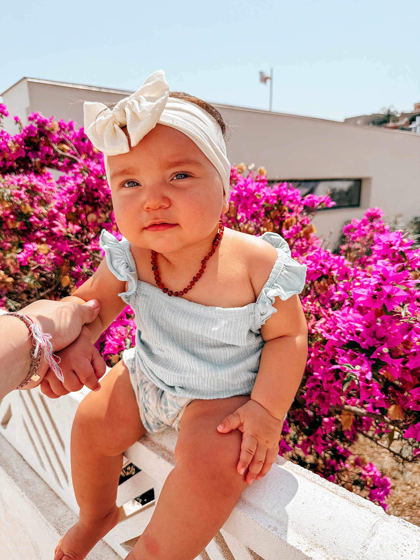 Kelya a rejoint le concours — aidez-le/la à gagner de superbes lots ! baby, child, headband, bow, necklace, hand, flower, pink_flower, outdoor, sunlight, wall, blue_sky, person, summer_clothing, cute, portrait, skin, holding_hand, nature, happy