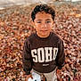 child, boy, autumn, fall_leaves, pumpkin, sweatshirt, curly_hair, outdoor, casual_clothing, standing, nature, seasonal, leaf_litter, portrait, young, person, daylight, brown, white, shoes