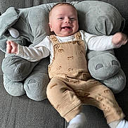 Malo a rejoint le concours — aidez-le/la à gagner de superbes lots ! baby, infant, smiling, overalls, plush_elephant, couch, grey_fabric, happy, child, soft_toy, cute, kicking, sitting, indoor, clothing, face, person, young_child, home, comfort
