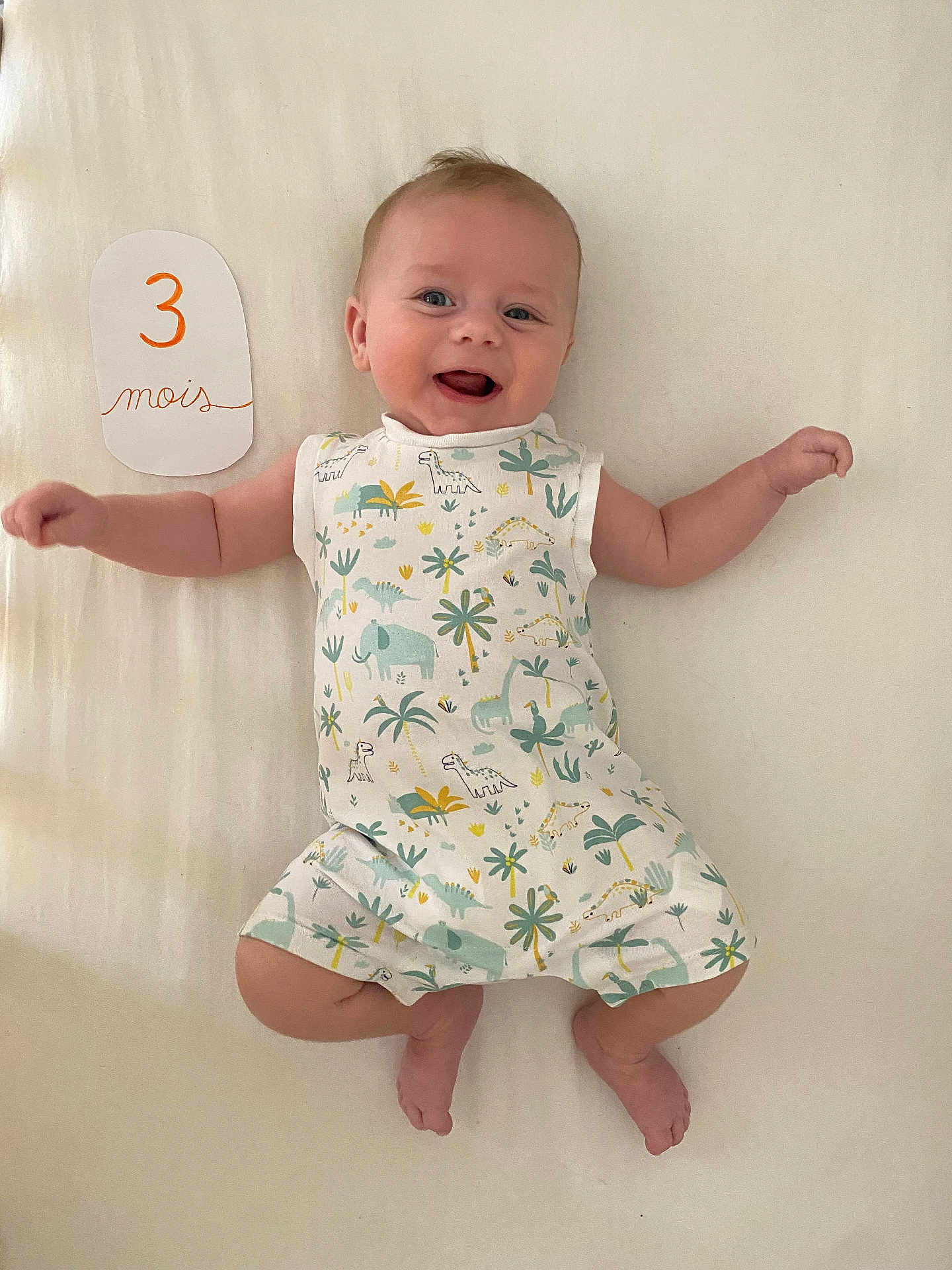 Malo participe au concours pour gagner de l'argent avec cette photo : baby, infant, smiling, lying_down, romper, dinosaur_print, arms_outstretched, handwritten_sign, cream_background, cute, happy, child, one_person, indoors, closeup, skin, feet, face, blue_eyes, light