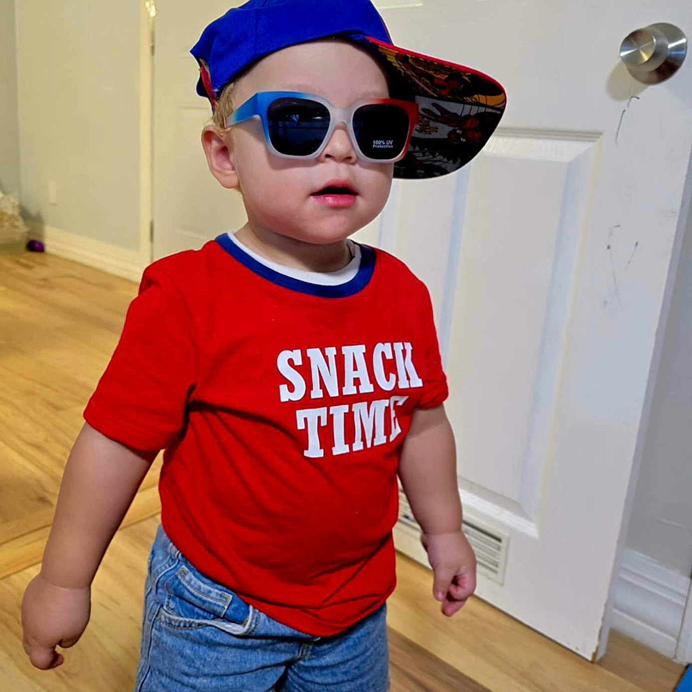 Liam is registered to the contest to win money with this photo: blue_cap, boy, casual, child, cute, denim_shorts, door, fashion, hat, indoor, portrait, red_shirt, smiling, snack_time, socks, standing, sunglasses, toddler, white_door, wooden_floor