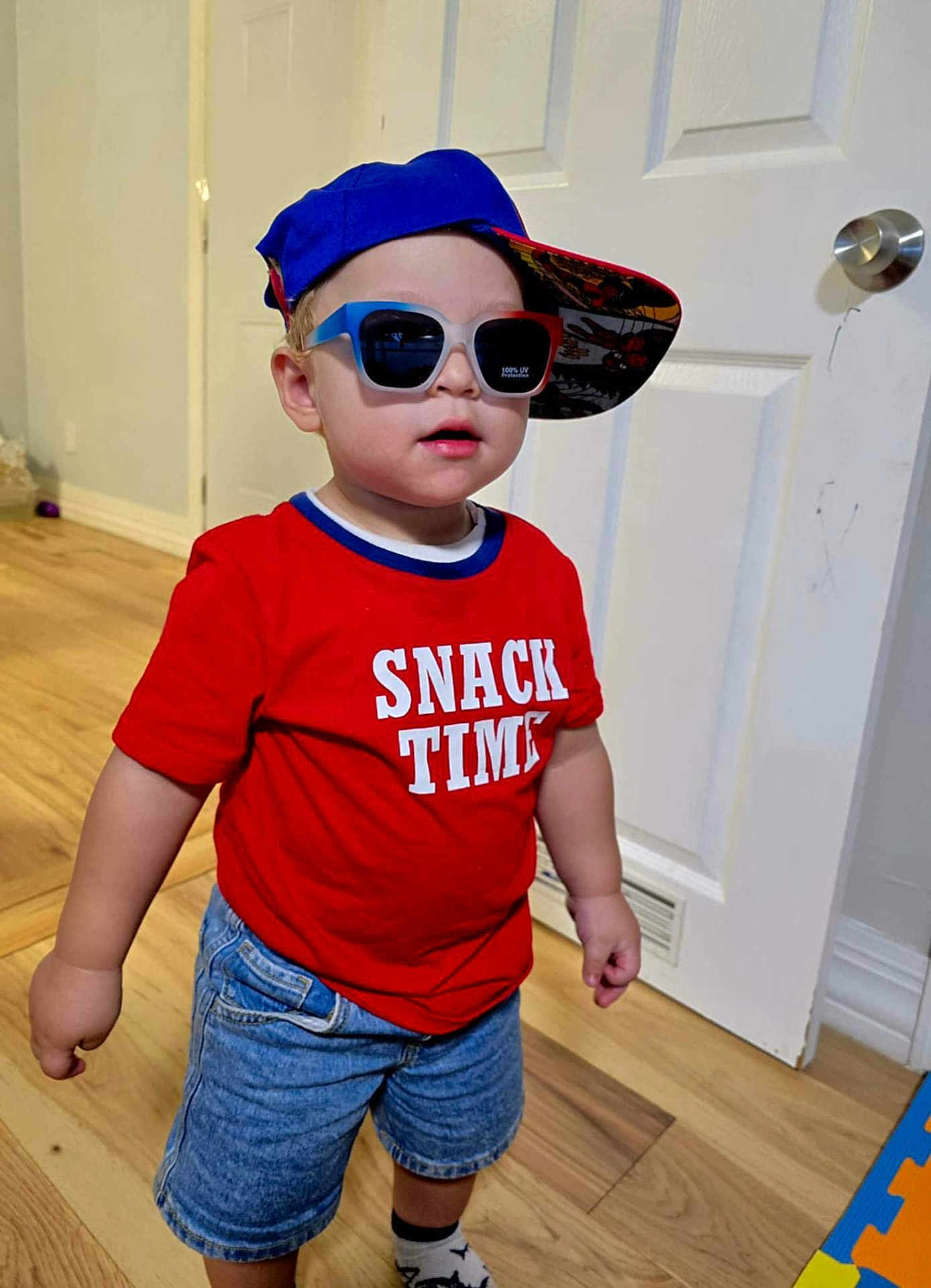 Liam is registered to the contest to win money with this photo: toddler, child, boy, red_shirt, snack_time, blue_cap, sunglasses, denim_shorts, socks, indoor, wooden_floor, door, white_door, standing, fashion, casual, cute, portrait, smiling, hat