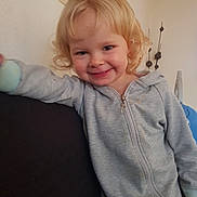 Emma participe au concours pour gagner de l'argent avec cette photo : child, smiling, blonde_hair, curly_hair, grey_jumpsuit, zipper, indoor, couch, leaning, happy, face, person, young_child, wall_decor, home, casual, light_skin, cozy, portrait, toddler