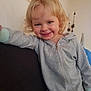 child, smiling, blonde_hair, curly_hair, grey_jumpsuit, zipper, indoor, couch, leaning, happy, face, person, young_child, wall_decor, home, casual, light_skin, cozy, portrait, toddler
