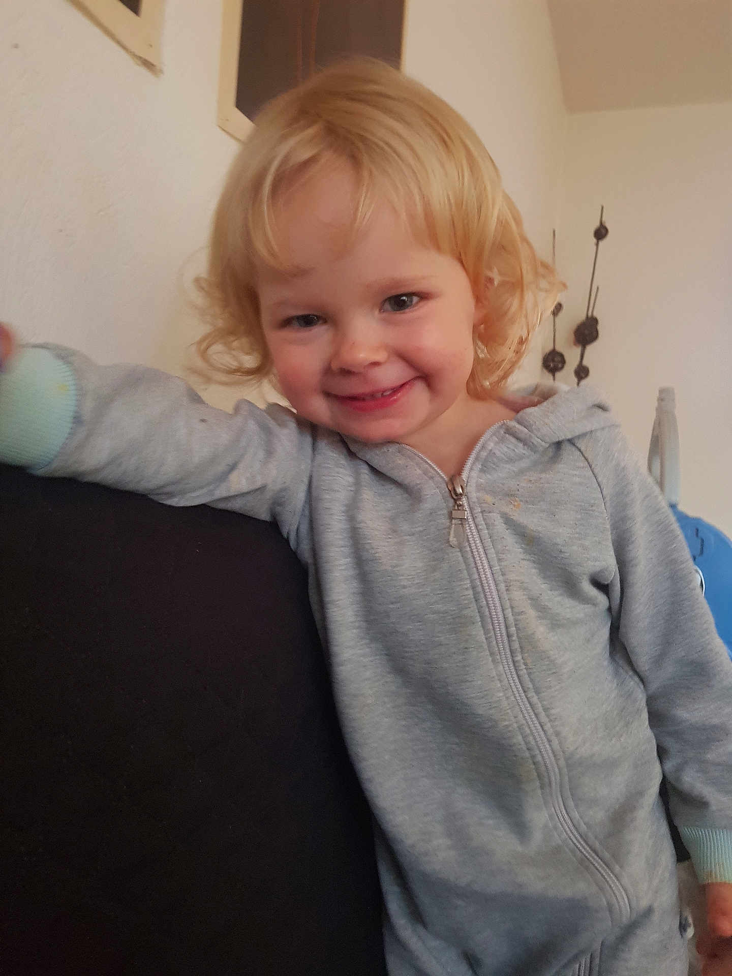 Emma participe au concours pour gagner de l'argent avec cette photo : child, smiling, blonde_hair, curly_hair, grey_jumpsuit, zipper, indoor, couch, leaning, happy, face, person, young_child, wall_decor, home, casual, light_skin, cozy, portrait, toddler
