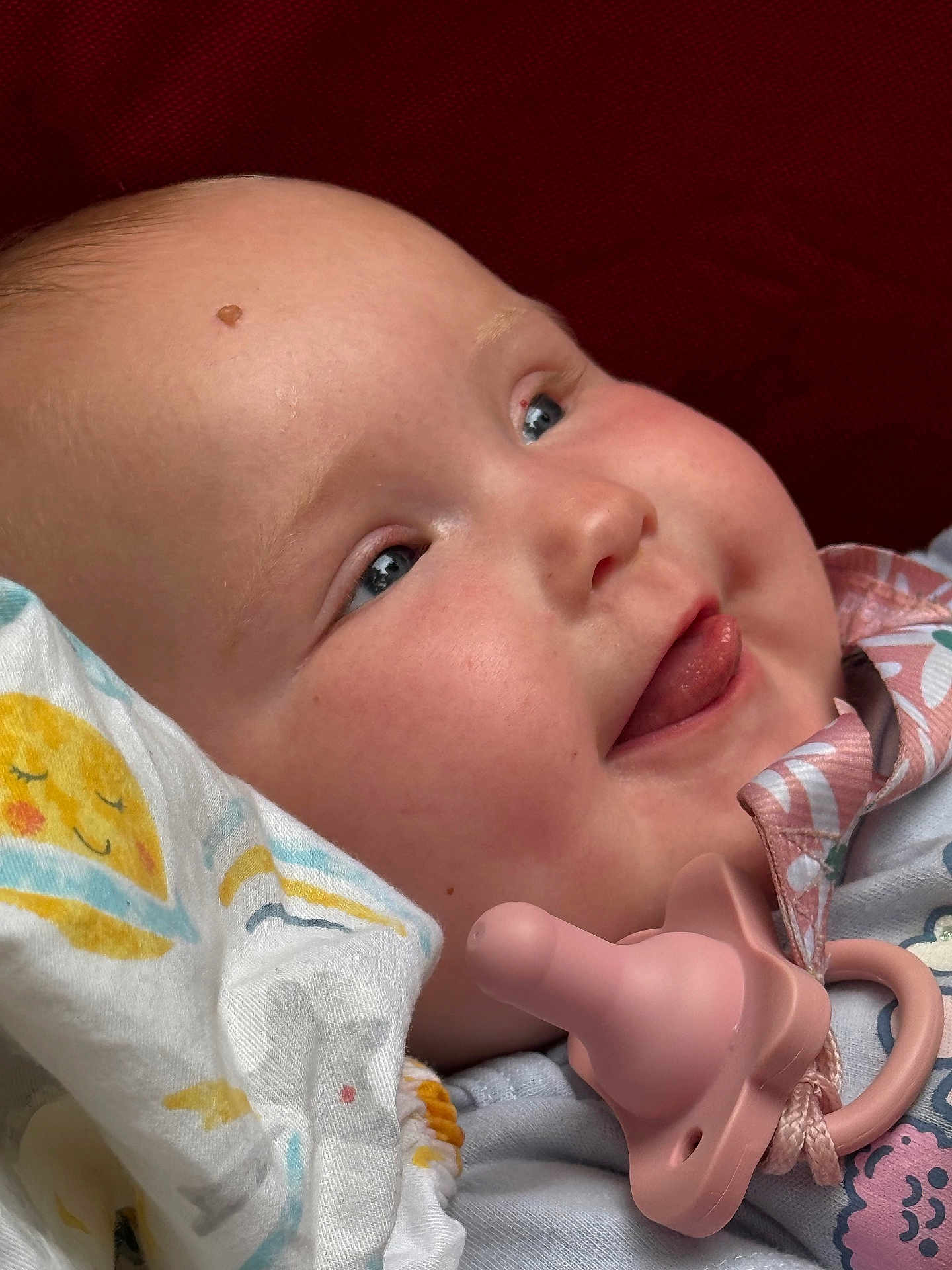 Freya is registered to the contest to win money with this photo: baby, face, tongue, pacifier, blanket, blue_eyes, close_up, smile, infant, clothing, pink, patterned, soft, cute, cheeks, skin, head, lying_down, happy, child